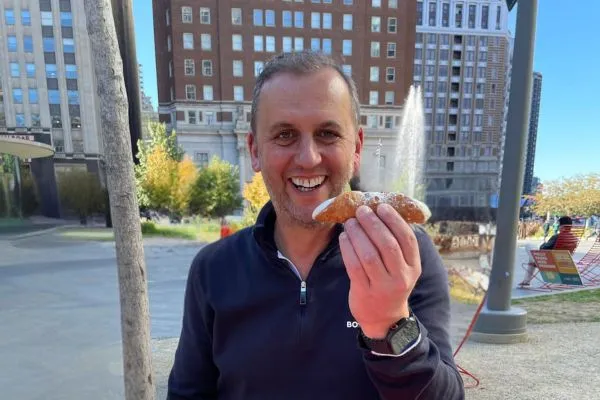 Mark Turnbull sitting in Philadelphia, smiling and holding a Cannoli during a trip to meet fellow independent retailers.