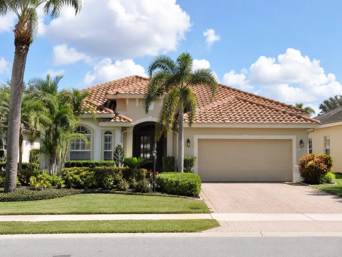 Florida suburban home with a terracotta tile roof, palm trees, tropical landscaping, and a paved driveway under a bright blue sky.
