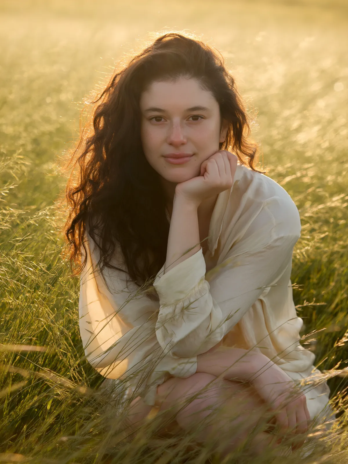 Barbara Guimarães sitting in a sunlit field, wearing a soft cream blouse and looking gently at the camera; warm, serene portrait reflecting Mental Nesting’s gentle healing aesthetic.