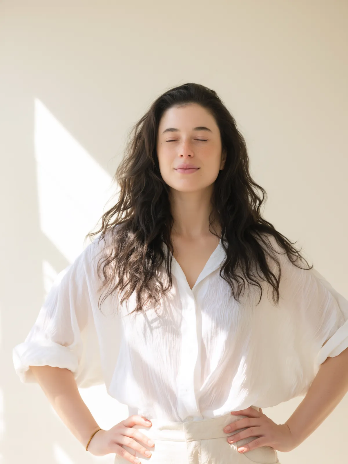 Barbara Guimarães standing in natural light with eyes closed and hands on hips, wearing a white blouse; peaceful, grounded expression representing nervous system regulation and inner healing.