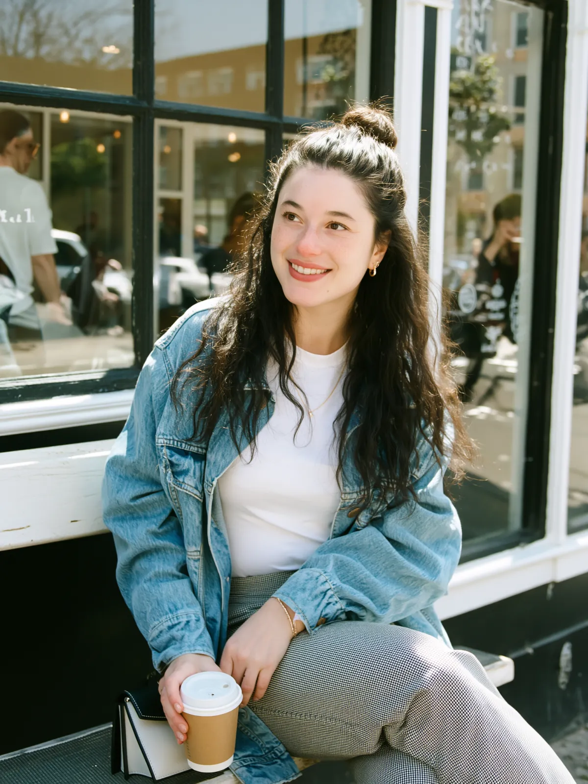 Barbara Guimarães sitting on a bench outside a coffee shop, wearing a denim jacket and holding a cup of coffee while looking off to the side with a soft smile.