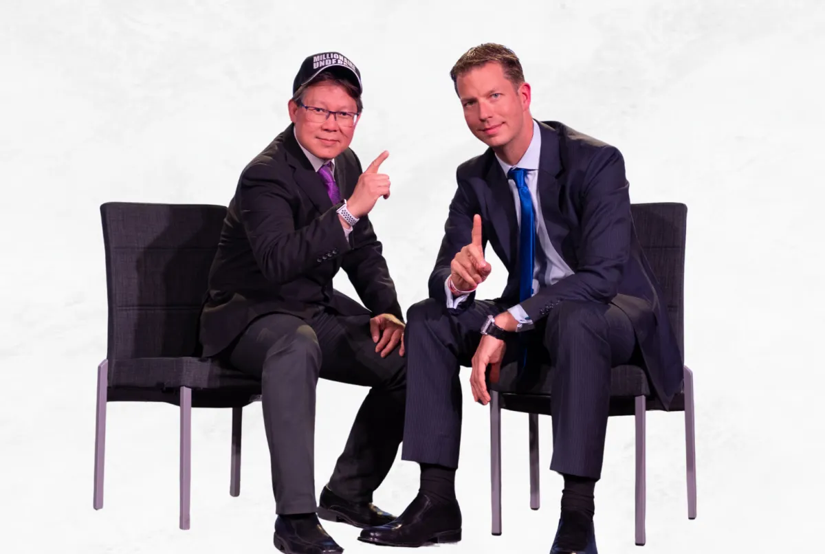 Sadikin Budiman seated with JT Foxx on stage, both in suits, posing for a photo during an event.