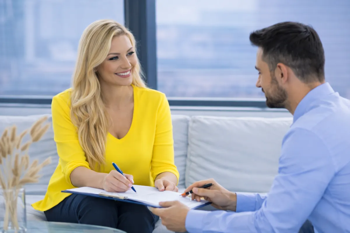 Two professionals seated on a sofa in a modern office, smiling and discussing documents on a clipboard, with large windows and city views in the background.