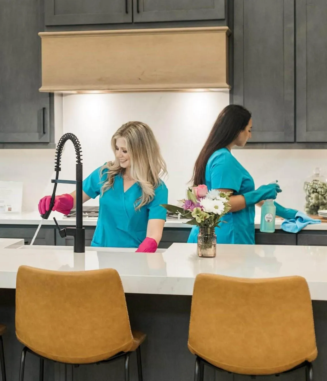 Two professional cleaners from **True Cleaning Experts** are thoroughly cleaning a modern kitchen. One cleaner, wearing bright pink gloves, is wiping down the sink and faucet, while the other, wearing blue gloves, is disinfecting the countertop with a spray bottle and cloth. Both are dressed in matching blue uniforms, showcasing their professionalism. The kitchen features sleek cabinetry, elegant countertops, and decorative touches like a flower arrangement, reflecting the team's dedication to achieving a spotless and welcoming environment.