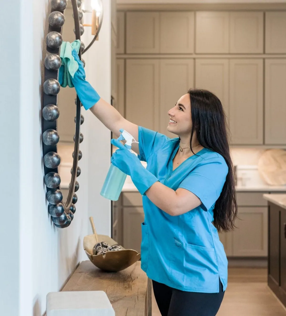 A professional cleaner from **True Cleaning Experts** is smiling as she cleans a decorative mirror in a stylish, modern home. She is wearing a blue uniform and protective cleaning gloves, holding a spray bottle in one hand and a microfiber cloth in the other. The clean and organized space features wooden furniture and a neatly designed interior, reflecting the team's commitment to delivering sparkling, spotless results.