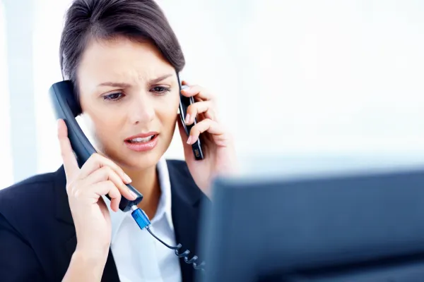 Stressed businesswoman answering multiple phone calls while working at a computer, representing slow lead response, missed calls, and overwhelmed customer inquiries