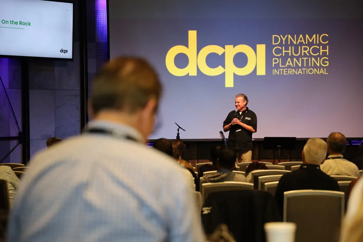 A speaker stands on a stage holding a microphone, addressing an audience seated in rows of chairs. The large screen behind him displays the logo "DCPI" along with the words "Dynamic Church Planting International." The speaker is wearing a black shirt with the DCPI logo and is smiling while speaking. The setting appears to be a conference or seminar focused on church planting. The lighting in the room is professional, with purple and blue hues on the walls, and a guitar is visible on the stage. The audience is attentively listening, with one person in the foreground slightly blurred.