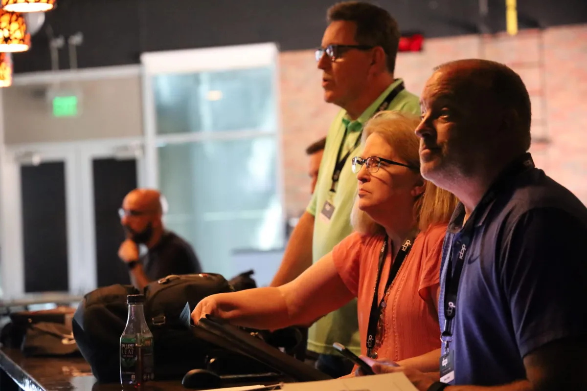 A group of three people, two men and one woman, are attentively watching something off-camera. They are standing at a counter or table, with bags and a bottle of tea visible in the foreground. The lighting is dim, and the background is slightly blurred, giving the image a focused and contemplative atmosphere. The individuals are wearing lanyards, suggesting they are at an event or workshop. The scene suggests a moment of concentration or discussion.