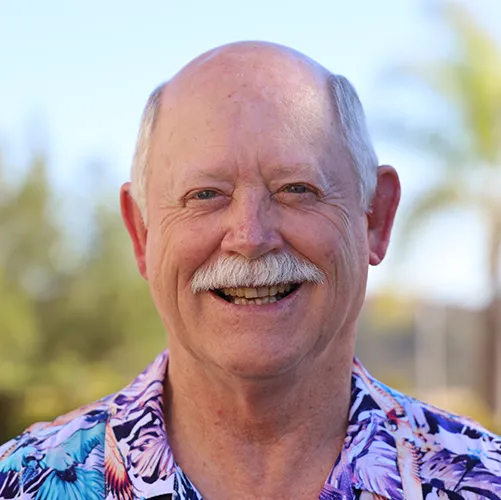 Smiling older man with a bald head and mustache, wearing a colorful tropical shirt, standing outdoors with trees and foliage in the background