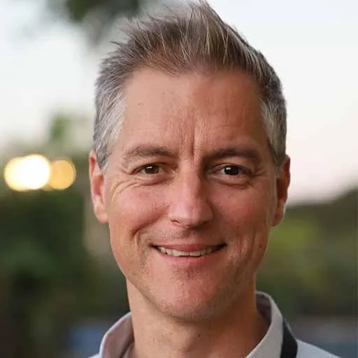 Smiling man with short, light-colored hair, wearing a light shirt with a dark strap over his shoulder, standing outdoors with a blurred background of trees and lights