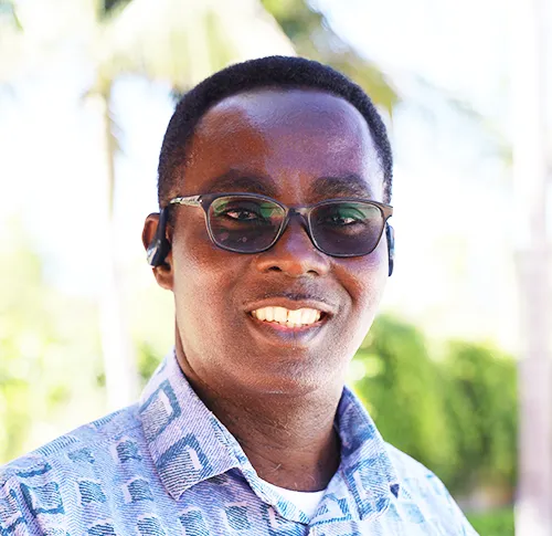 Smiling man wearing glasses and a patterned shirt, standing outdoors with trees and foliage in the background