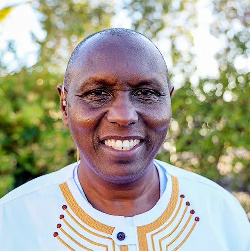 A smiling man with short-cropped hair, wearing a white shirt with yellow and red embroidery, standing outdoors with greenery in the background