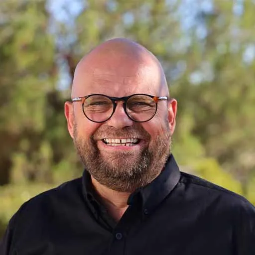 A smiling man with glasses, a bald head, and a beard, wearing a black shirt, standing outdoors with greenery in the background