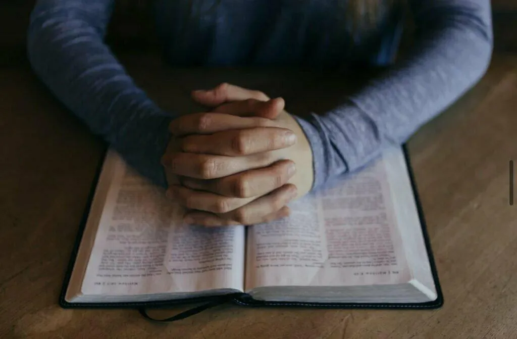 Open Bible on a table with folded hands n top of it. You can see part of the arms in the background.