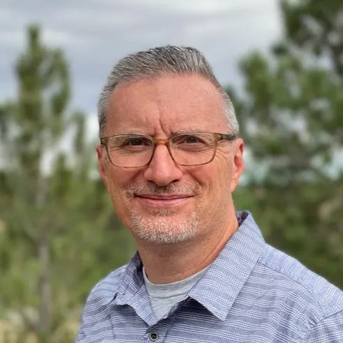 Smiling man with short gray hair, glasses, and a trimmed beard, wearing a light blue checked shirt, standing outdoors with trees in the background