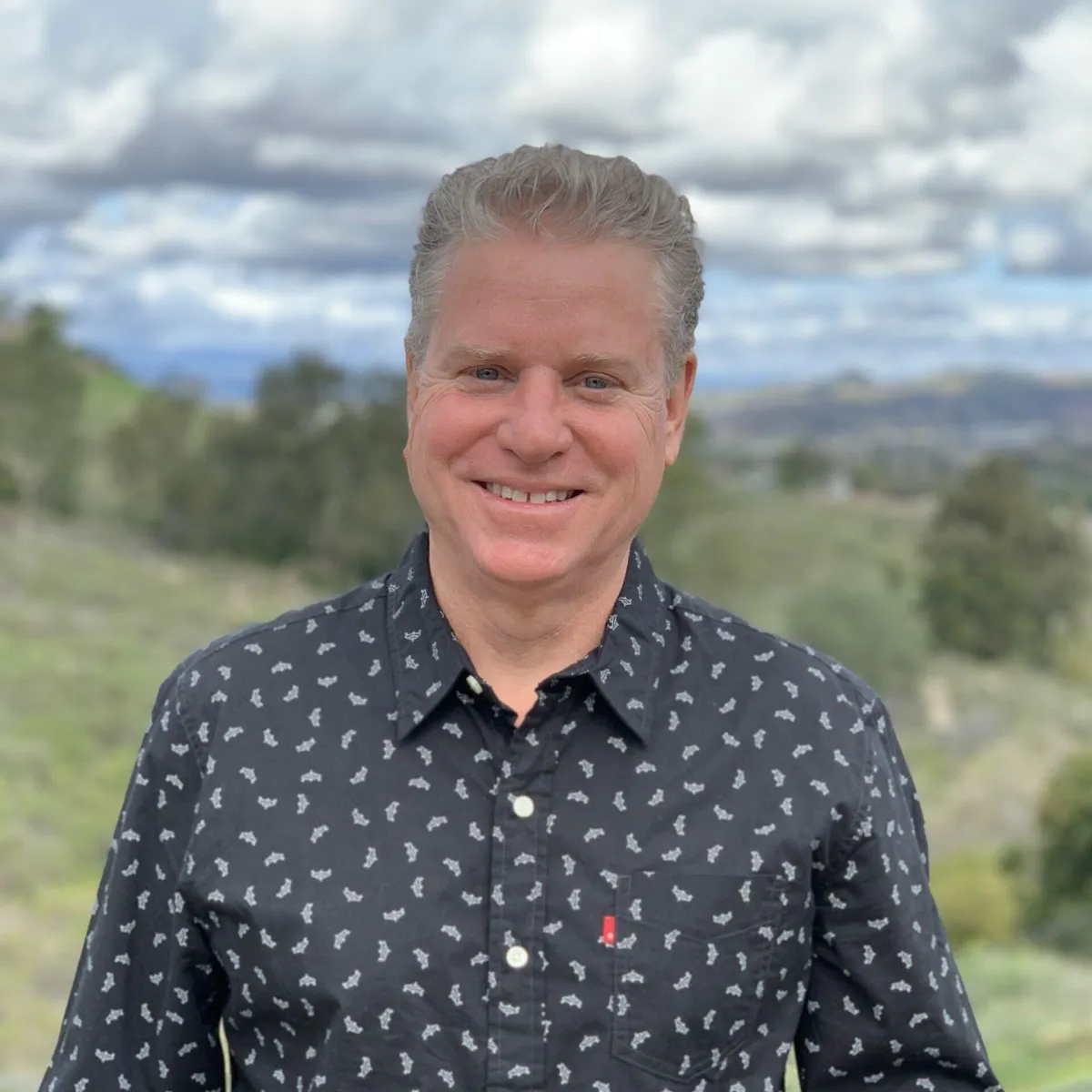 Smiling man with short, light-colored hair, wearing a dark patterned shirt, standing outdoors with a scenic background of hills, trees, and a cloudy sky.