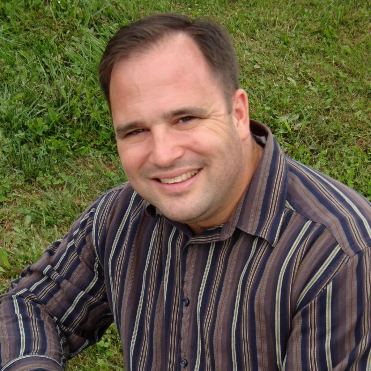 Smiling man with short dark hair, wearing a striped shirt, sitting on the grass outdoors.