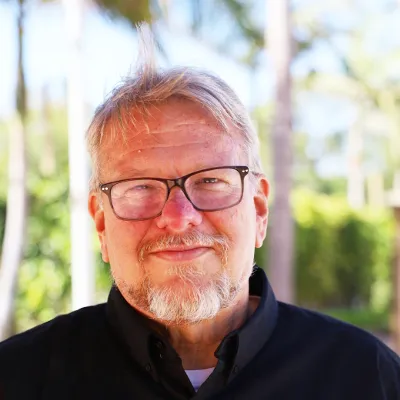 Smiling older man with light hair, beard, and glasses, wearing a black shirt, standing outdoors with trees and foliage in the background