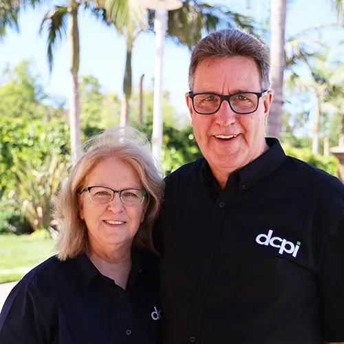 A smiling man and woman standing outdoors, both wearing black shirts with the 'dcpi' logo. The man has short brown hair and glasses, and the woman has shoulder-length blonde hair and glasses. They are standing in front of a background featuring palm trees and greenery.