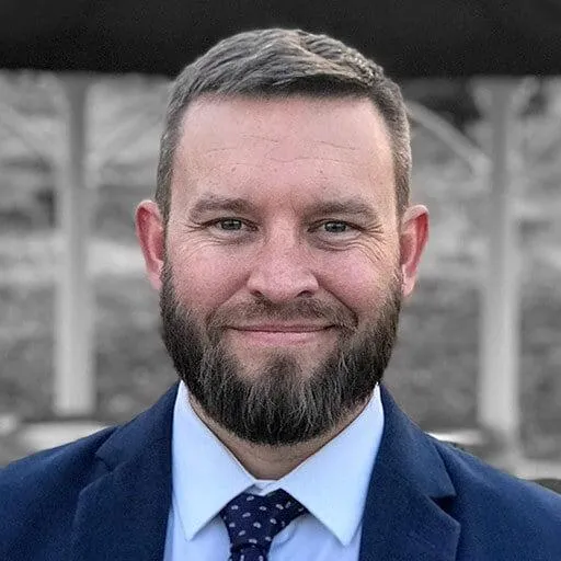 A portrait of a man with a neatly trimmed beard and short hair, wearing a dark blue suit, white shirt, and patterned tie. He is smiling slightly and looking directly at the camera. The background is blurred, showing an outdoor setting with muted colors, possibly a park or garden area.