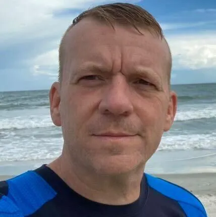 Middle-aged man standing on a beach with the ocean and sky in the background, wearing a blue and black athletic shirt, with short, light brown hair and a serious expression.