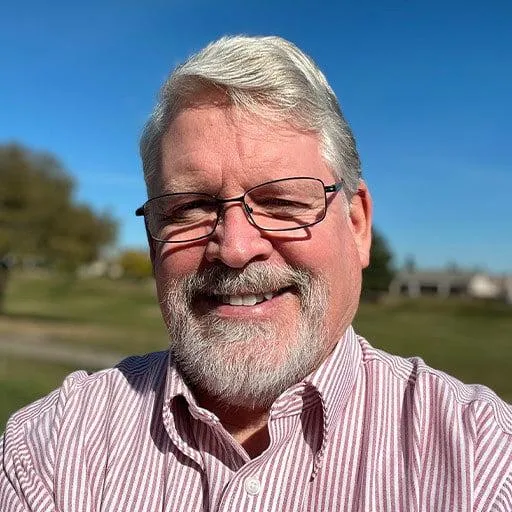 A man with gray hair and a beard is smiling at the camera. He is wearing glasses and a striped button-up shirt. The background features a clear blue sky and a grassy outdoor area with trees, suggesting a pleasant, sunny day. His expression is warm and friendly.