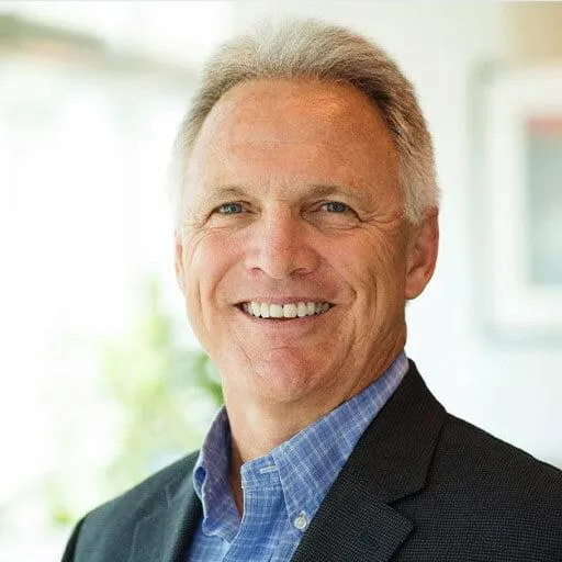 A portrait of a smiling man with short gray hair, wearing a blue checkered shirt under a dark blazer. He has a friendly and approachable expression, with a blurred background that suggests a professional setting.