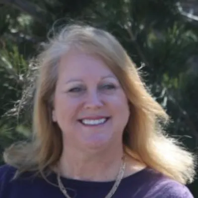 Middle-aged woman with long blonde hair, wearing a purple top and a gold necklace, smiling warmly with greenery in the blurred background.