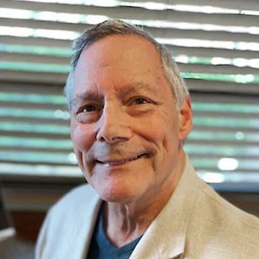 Older man with gray hair, wearing a beige suit jacket over a dark shirt, smiling warmly with a blurred background of horizontal blinds and indoor elements.