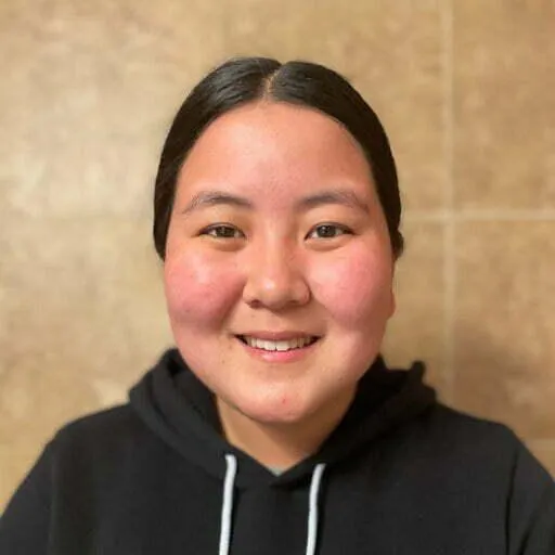 A portrait of a young woman with straight black hair pulled back, smiling softly at the camera. She is wearing a black hoodie, and the background is a plain beige or tan wall, giving a warm and neutral backdrop to the image.