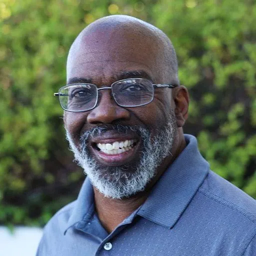 A portrait of a man with a bald head and a full gray beard, smiling broadly at the camera. He is wearing glasses and a light blue polo shirt. The background is outdoors, with green foliage providing a natural and vibrant backdrop.