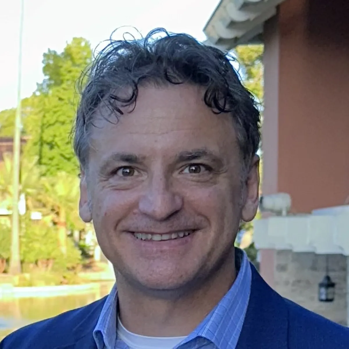 Middle-aged man with curly dark hair, wearing a blue blazer over a light blue shirt, smiling warmly with an outdoor background featuring trees and a building.