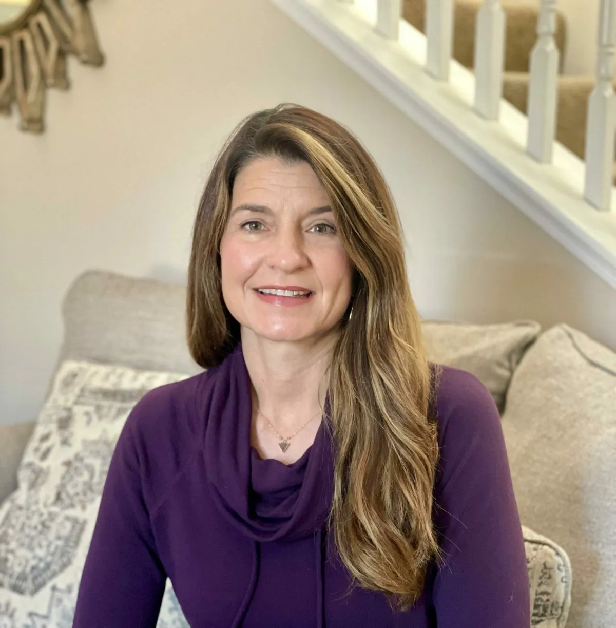 A woman with long brown hair, wearing a purple sweater, sits on a couch in a cozy living room. She is smiling softly at the camera. Behind her, there is a staircase and a patterned cushion, adding warmth and comfort to the scene.