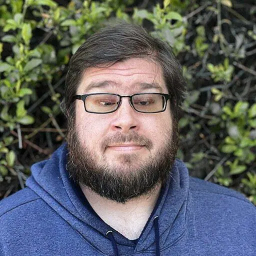 A man with a full beard and glasses, wearing a blue hoodie, stands outdoors with greenery in the background. He has a thoughtful expression on his face as he looks slightly downward.