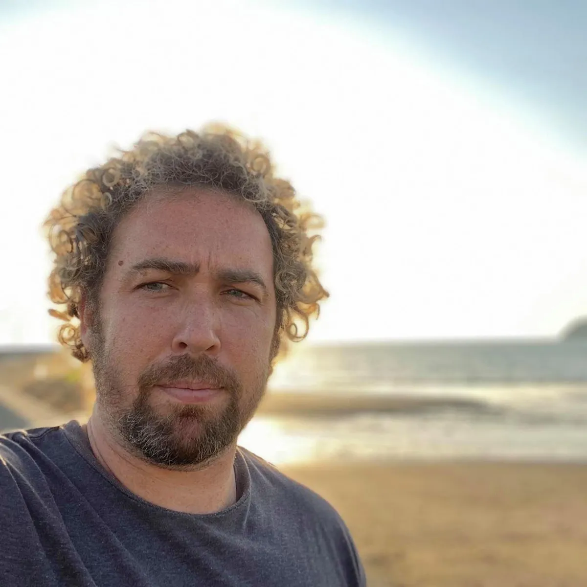 Man with curly hair and a beard, wearing a dark gray shirt, standing on a beach at sunset with the ocean and a distant shoreline in the background, looking directly at the camera with a serious expression.