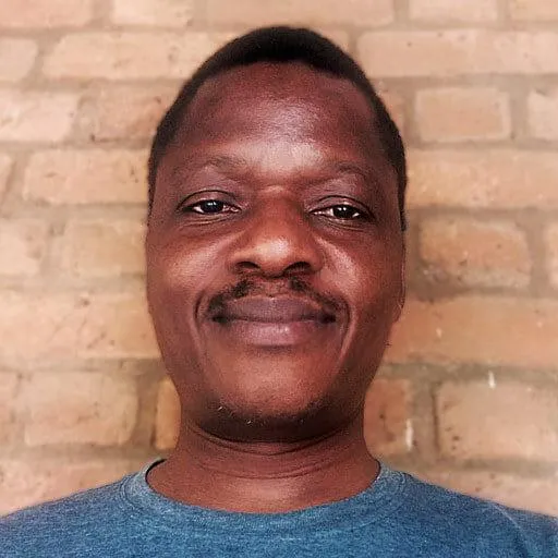 A man with short hair and a slight mustache, wearing a blue t-shirt, stands in front of a brick wall. He is smiling gently and looking directly at the camera. The brick wall in the background is slightly blurred, drawing focus to the man's face.