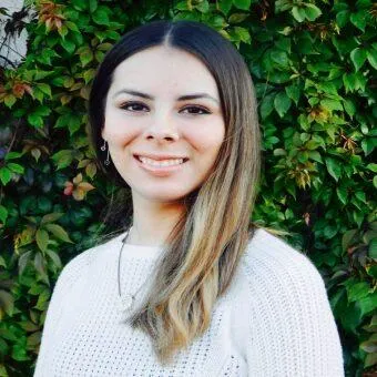 Young woman with long dark hair and blonde highlights, wearing a white sweater and earrings, smiling warmly with a background of green leafy vines.