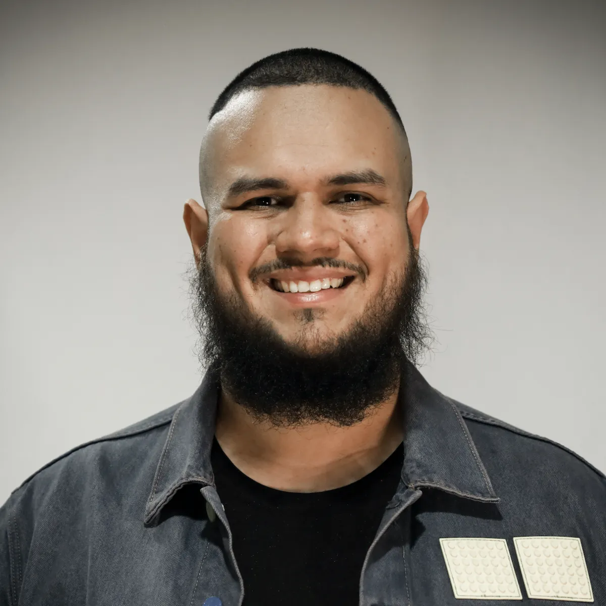 Man with a shaved head and a thick beard, wearing a dark denim jacket over a black shirt, smiling warmly against a plain background.