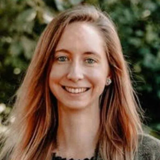 A woman with long, reddish-blonde hair is smiling at the camera. She is wearing a dark green top and standing outdoors with greenery in the background. Her expression is friendly and cheerful, conveying a warm and approachable demeanor.