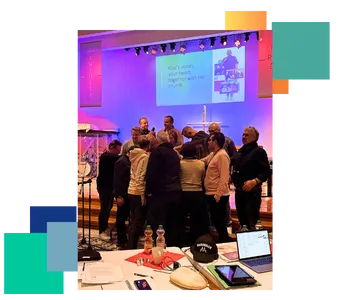 A group of people standing in a circle at the front of a stage, possibly in prayer or a discussion, under purple and blue lighting. Behind them, a screen displays the text 'God’s vision, your heart, together with His church.' In the foreground, there are tables with notebooks, laptops, and bottles, suggesting a workshop or conference setting.