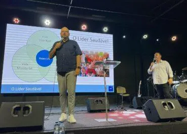 Two men are standing on a stage, one holding a microphone and speaking. Behind them is a large screen displaying a presentation titled 'O Líder Saudável' with a Venn diagram and images of people. The stage is set up with audio equipment, and the room has dim lighting.