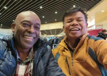 Two men smiling for a selfie in what appears to be an indoor public space with modern ceiling design and warm lighting. Both are wearing jackets, and they look happy and relaxed.