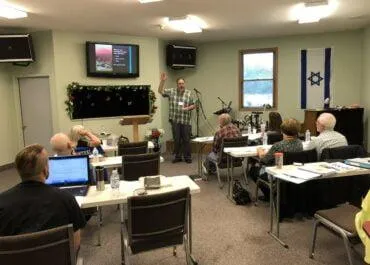 A man standing in front of a classroom, giving a presentation to a group of seated attendees. The presentation is projected on a screen, and the speaker is holding a microphone. There are laptops and notebooks on the tables, and a large Israeli flag hangs on the wall. The room has a casual setup with microphones and instruments in the background.