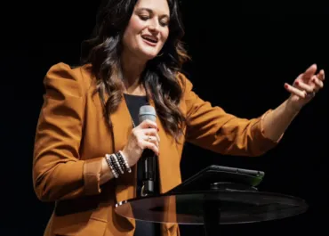 A woman wearing a brown blazer is speaking into a microphone while gesturing with her hand. She is standing behind a podium with a tablet placed on it, delivering a presentation or speech. The background is dark, focusing attention on the speaker.