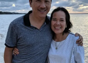 A couple smiling and posing for a photo near the water at sunset. The man is wearing a gray polo shirt, and the woman is wearing a white, polka-dotted blouse. They have their arms around each other, and the sky in the background is cloudy, with soft evening light.