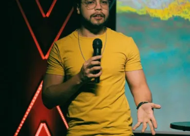 A man wearing a yellow t-shirt and glasses is holding a microphone while speaking on stage. He is gesturing with one hand, and behind him is a screen displaying colorful visuals. The stage is decorated with illuminated red accents.