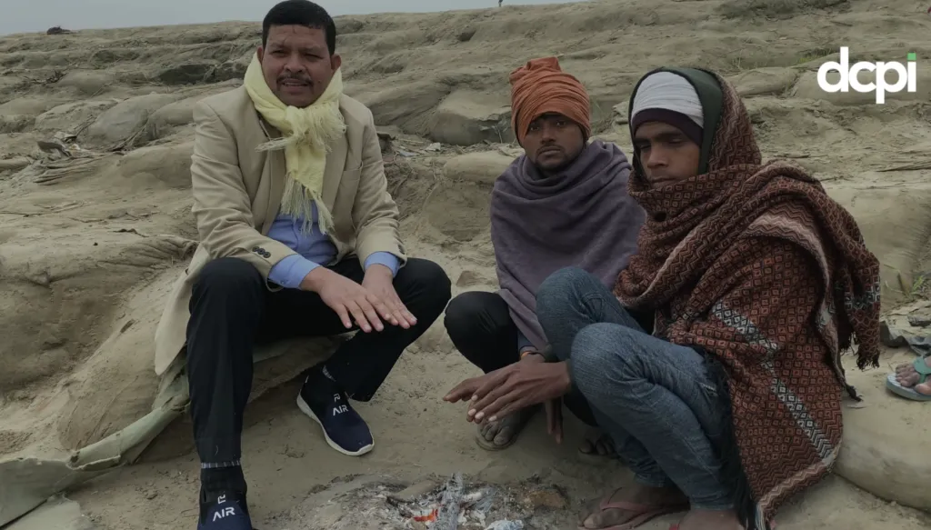 Three men sitting on a sandy, barren landscape around a small fire. One man, dressed in a beige coat and scarf, is on the left, while the other two men are wrapped in shawls and head coverings. They appear to be warming their hands by the fire. The logo 'dcpi' is visible in the top right corner of the image.