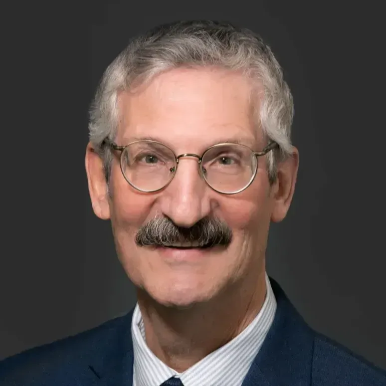 A circular photo of a middle-aged man with gray hair and a mustache, wearing glasses, a beige blazer, and a patterned tie. He is seated at a desk, smiling and gesturing with his hands, likely in the middle of a conversation or presentation. The background shows part of a whiteboard with some writing on it, suggesting an office or meeting room environment.
