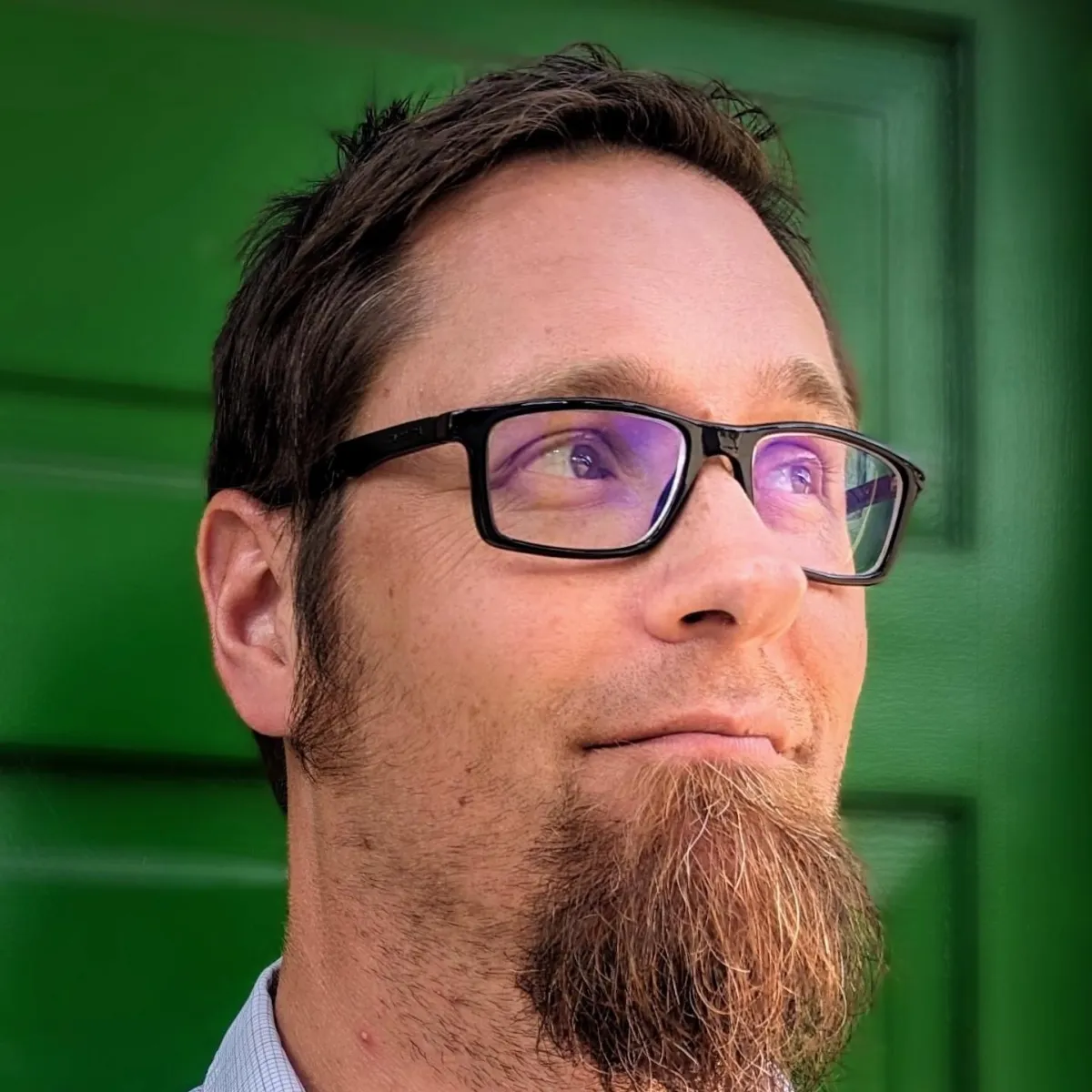 A man with a full beard and glasses, wearing a blue hoodie, stands outdoors with greenery in the background. He has a thoughtful expression on his face as he looks slightly downward.