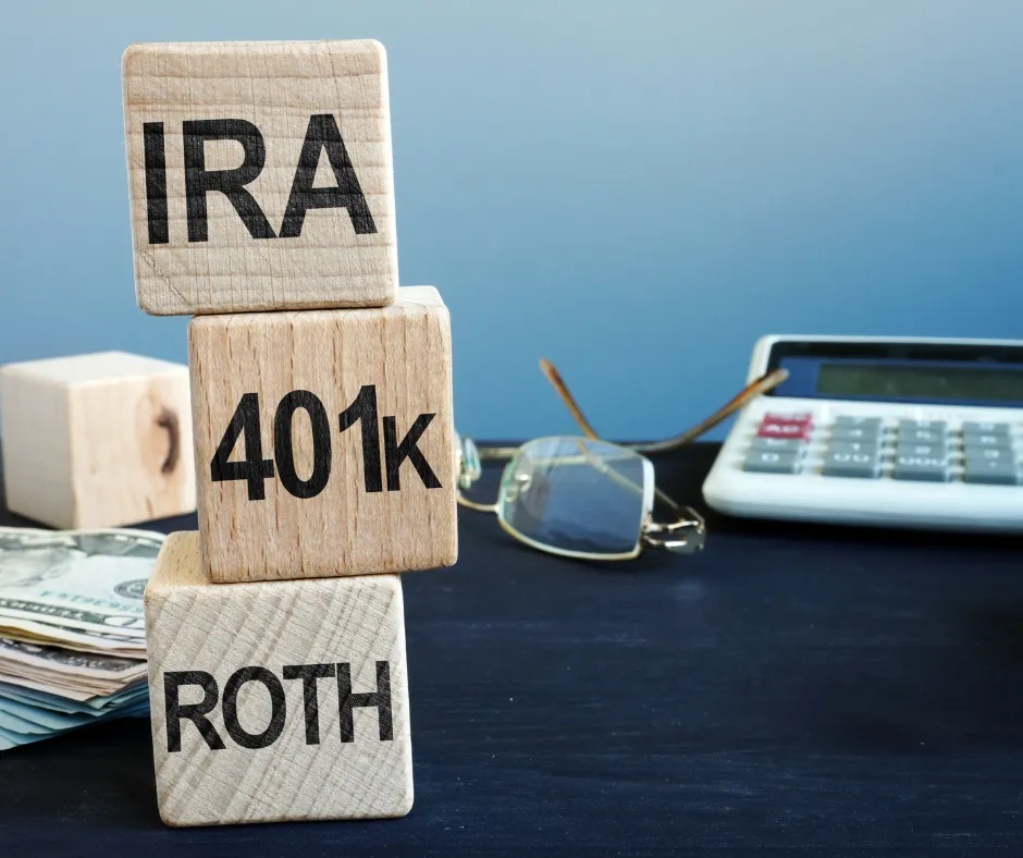 Wooden blocks labeled IRA, 401k, and Roth stacked on a desk with cash, a calculator, and glasses in the background, symbolizing retirement savings and financial planning.